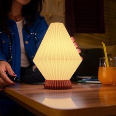 a woman sitting at a table with a lamp on it's side and an orange drink in front of her