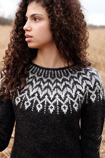 a woman standing in a wheat field wearing a black sweater with white and gray designs