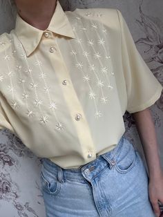 This vintage blouse is made from a delicate pale yellow fabric, featuring short sleeves and adorned with intricate embroidery. The feminine design is highlighted by elegant pearl buttons that add a touch of sophistication. Ideal for both casual and dressy occasions, this shirt embodies the timeless charm of vintage fashion with its detailed craftsmanship and classic silhouette. - Good vintage condition  - TAG SIZE: 44 - Fabric information: 100% polyester  Estimated to fit XS-S-M-L based on your Elegant Half Sleeve Summer Shirt, Cream Short Sleeve Blouse For Summer, Elegant Beige Summer Shirt, Feminine Short Sleeve Cream Blouse, Feminine Cream Short Sleeve Blouse, Elegant Yellow Summer Shirt, Beige Formal Summer Shirt, Beige Formal Shirt For Summer, Elegant Embroidered Summer Shirt