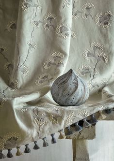 a vase sitting on top of a wooden table next to a white curtain with tassels