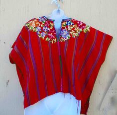 "A wide all cotton hand made vintage blouse or huipil that is brilliant bright red all over, with a border of hand-embroidered flowers and leaves at neck area of blouse, both front and back. Sleeves are short, wide at shoulders, has ten vertical embroidered rainbow ( multicolored) travel down top, each speed 2\" apart. Center line has a band of blue, pink, and green embroidery running down to bottom. Top is hand-sewn up each side with bright green thread, and bottom hem is a rough extra panel as Bohemian Red Blouse With Traditional Patterns, Red Bohemian Blouse With Traditional Patterns, Red Embroidered Blouse For Festivals, Red Blouse With Embroidered Border For Festivals, Traditional Red Blouse For Summer, Traditional Red Blouse With Embroidered Border, Traditional Red Blouse With Traditional Patterns, Traditional Red Tops For Festival, Red Blouse With Traditional Patterns For Festivals