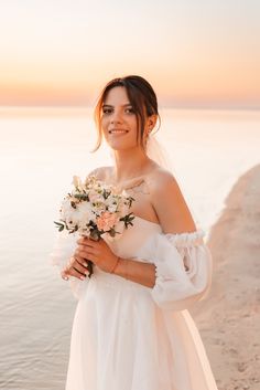 a woman in a wedding dress holding a bouquet of flowers on the beach at sunset