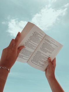 two hands hold open a book in front of a blue sky with white fluffy clouds