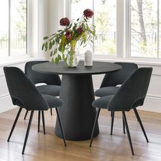 a round table with four chairs around it and a vase on the top, in front of two windows