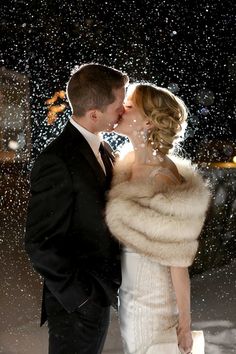 a bride and groom kissing in the rain at their wedding reception, with snow falling all around them