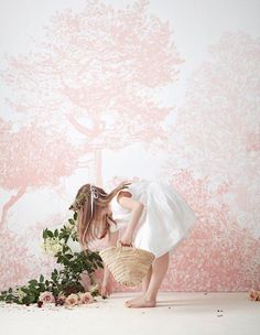 a woman bending over holding a basket with flowers on it in front of a pink wall