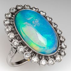 This gorgeous ring is centered with one (1) oval cabochon cut natural crystal opal and set into a bezel setting. The opal is bordered with twenty-two (22), bezel set, round brilliant cut diamonds, The top face of the shank is accented with three (3), bead set, round brilliant cut diamonds on each side. The gemstones are bordered with milgrain edging. The ring measures 29.1mm at the top, rises 10.6mm above the finger, tapering to 1.8mm wide and 1.1mm thick at the base of the shank. This ring is currently a size 6. Oval Cabochon Opal Ring Fine Jewelry, Fine Jewelry Oval Cabochon Opal Ring, Oval Cabochon Opal Ring In Fine Jewelry, Exquisite Oval Multi-stone Opal Ring, Exquisite Multi-stone Oval Opal Ring, Opal Ring With Diamond Accents For Formal Occasions, Oval Opal Ring With Bezel Setting For Formal Occasions, Formal Oval Opal Ring With Gemstone Accents, Oval Cabochon Diamond Opal Ring