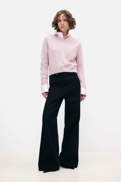 a woman standing in front of a white background wearing black pants and a pink sweater