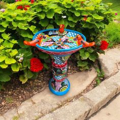 a colorful bird bath sitting in the middle of some flowers and bushes with green leaves