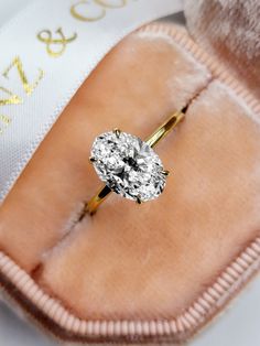 an oval diamond ring sits on top of a pink velvet case with a white ribbon around it