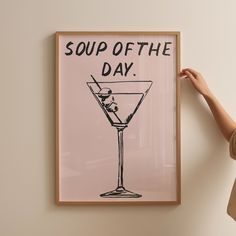 a woman holding up a framed poster with a martini in it that says soup of the day