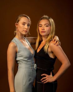 two women standing next to each other in front of a brown background, one wearing a black dress