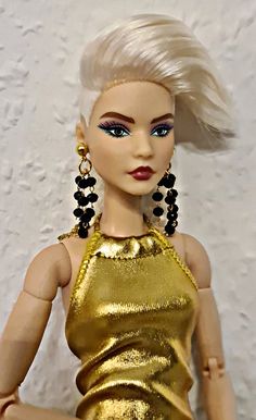 a doll with blonde hair and blue eyes wearing a gold dress, black beaded earrings