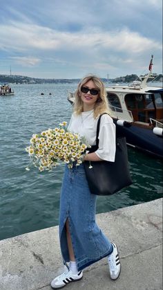 a woman is walking by the water with flowers in her hand and holding a black bag