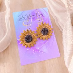 a pair of sunflower earrings sitting on top of a purple card next to white lace