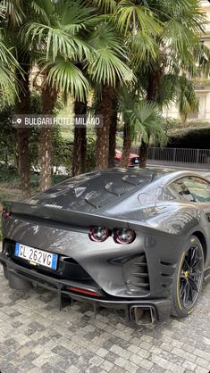 a grey sports car parked in front of some palm trees and bushes on a brick sidewalk