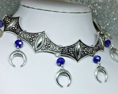 Victorian Gothic metal choker with crescent moon charms and blue crystal beads, ornate metal necklace, Victorian jewelry, Gothic jewelry A beautifully ornate gothic silver metal choker with Victorian crescent moon charms and blue faceted crystal beads. It measures approx. 11 inches in length and has lobster clasp fastening and an extension chain, so that it can be adjusted to fit the neck. The choker is made up of 7 silver toned metal ancient style sections that have intricate detailing. A marve Bohemian Crescent Metal Necklaces, Gothic Blue Metal Jewelry, Crescent Metal Jewelry For Festivals, Metal Crescent Jewelry For Festivals, Blue Gothic Metal Jewelry, Blue Festival Jewelry With Moon Charm, Metal Crescent Shaped Festival Jewelry, Festival Blue Jewelry With Moon Charm, Crescent-shaped Metal Jewelry For Festivals