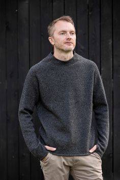 a man standing in front of a wooden wall wearing a gray sweater and khaki pants