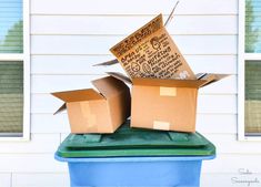 two cardboard boxes sitting on top of a trash can in front of a white house