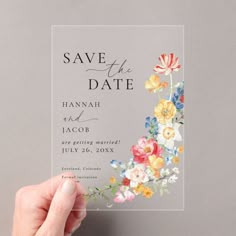 a person holding up a save the date card with colorful flowers on it in front of a gray background