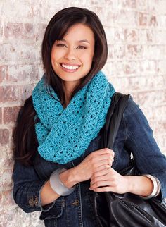 a woman wearing a blue crocheted scarf and carrying a black bag smiles at the camera