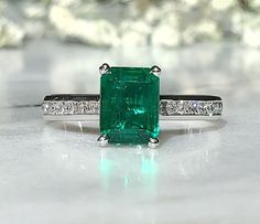 Simple, but modern emerald cut, emerald accented diamond ring. The center stone displays a gorgeous, Zambian mined emerald, set in a 4 double prong setting. The gallery shows off a round brilliant cut 1.6mm diamond. The sides of the shank have 20 1.7mm GHI, VS quality round diamonds. The ring shown in the photos is a 1.53ct Zambian stone. It is considered investment grade quality, which is the top quality of stone for an emerald. It has only been treated with minor colorless oil and the cut is a Luxury Emerald Ring With Baguette Cut And Accent Stones, Luxury Diamond Ring With Emerald Accent Stones, Modern Green Emerald Diamond Ring, Luxury Green Radiant Cut Rings, Luxury Asscher Cut Emerald Ring With Accent Stones, Modern Emerald Cut Emerald Ring With Accent Stones, Luxury Emerald Ring With Square Cut Diamond, Luxury Emerald Cut Emerald Ring In White Gold, Elegant Rectangular Emerald Ring Gia Certified