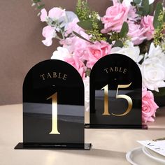 two black table numbers are sitting on a table with pink flowers in the back ground