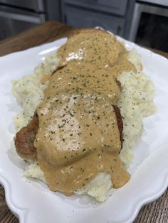 a white plate topped with meat covered in gravy and mashed potatoes on top of a wooden table