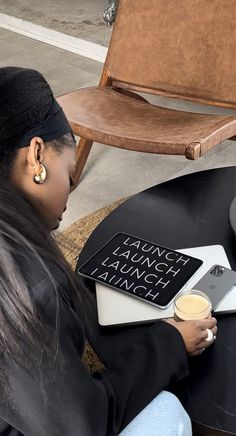 a woman sitting at a table with a laptop and coffee in her hand, reading a book