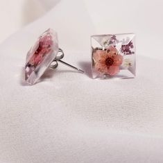 two square shaped earrings with flowers in them on a white cloth covered surface, one is pink and the other is purple