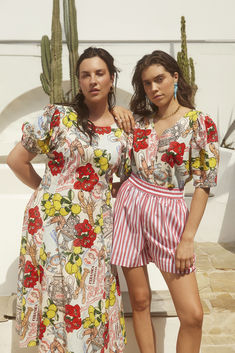 two women standing next to each other in front of a cactus and white building with red flowers on it