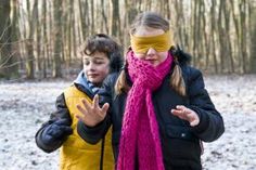 two children wearing blindfolds in the woods with one holding his face covered by a scarf