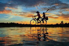 two people riding bikes across a body of water under a colorful sky with clouds in the background