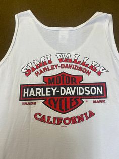 "Men's tank top Harley Davidson Moorpark Simi Valley, CA, 100% cotton, white color, size 3X. Good used condition, some stains and wear, check the pictures, official Harley Davidson product. Please, check carefully the measurements, photos and description of the article before buying it, we do not accept changes or returns. Measurements lying face down from outside: 28\" Across chest underarm to underarm. 15 1/2\" Shoulder seam to seam. 34\" Long, front middle top to bottom. Let me know if you ha White Letter Print Tank Top, White Sleeveless Tank Top With Letter Print, Basic White Cotton Tank Top, White Cotton Basic Tank Top, Casual White Tank Top, White Casual Lace Tank Top, White Racerback Top For Streetwear, White Sleeveless Cotton Muscle Tee, White Cotton Sleeveless Muscle Tee