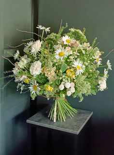 a bouquet of white and yellow flowers in a vase on a black stand against a green wall