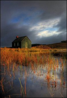 an old house sitting on top of a field next to a body of water with tall grass in front of it