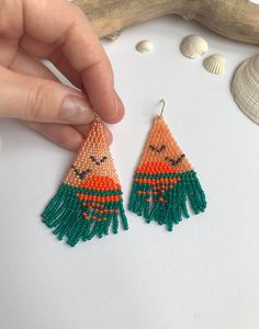 a pair of beaded earrings with orange and green tassels is being held by a person's hand