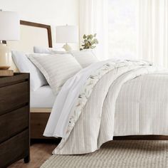 a bed with white comforter and pillows in a bedroom next to a lamp on a dresser