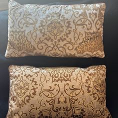 two decorative pillows sitting on top of a black couch in front of a window, one is gold and the other is brown