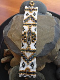 This hand-loomed bracelet is an original design that combines tiny rocaille seed beads with two sizes of larger, faceted Czech glass. The elegant white/black/gold color palette contains subtle shades of marbled picasso black, gun-metal gray, greystone, black diamond, and transparent topaz. The gold seed beads are plated in 24K gold. It fastens with a 24K gold vermeil toggle clasp. LENGTH: 7 inches WIDTH: 1.25 inches Artisan White Beaded Bracelets With Colorful Beads, Bohemian White Beaded Bracelets With Faceted Beads, Artisan White Beaded Bracelets, White Artisan Beaded Bracelets, Arrow Bracelet, Gold Color Palettes, Green Ombre, Loom Beading, Black Diamond