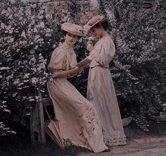 two women standing next to each other in front of bushes and flowers, one holding an umbrella