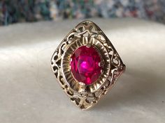 This stunning vintage ring is a beautiful example of antique jewelry craftsmanship, made of 10k gold and featuring a captivating, vibrant red faceted stone at its center. The stone, rich and intense in color, is securely set within an ornate setting that reflects a blend of late Victorian to Art Deco style, characterized by intricate lacy motifs and a delicate filigree pattern. The combination of the deep red stone and the intricate goldwork creates a striking visual contrast, enhancing the over Ornate Gold Ruby Ring, Antique Oval Red Ruby Ring, Red Filigree 14k Gold Jewelry, Red Victorian Filigree Ring With Gemstone, Victorian 14k Gold Ruby Ring In Red, Red 14k Gold Filigree Jewelry, Victorian Red Oval Filigree Ring, Antique Red Ruby Ring In 14k Gold, Victorian Ruby Ring In 14k Gold