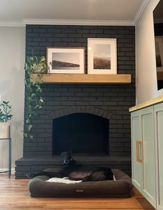 a dog is laying on his bed in front of the fireplace with two pictures above it