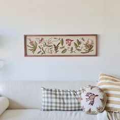 a white couch with pillows on it in front of a wall hanging above the couch