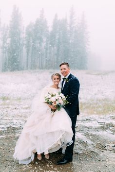 a bride and groom pose for a photo in the snow on their wedding day, with pine trees in the background