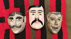 three masks with different facial expressions are on a red and black striped cloth, one has a moustache