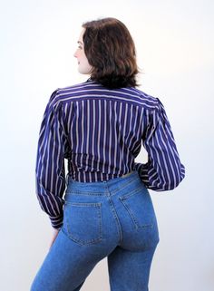 "This is a marvelous 1970s vintage blouse that has a lot going for it! The fabric is a silken poly with a very pleasant soft matte hand, like a charmeuse. The pattern is a classic vertical bar stripe in navy blue with taupe and a bright red pinstripe. It has shirring at the front shoulder yokes, puffed shoulders with small shoulder pads, long sleeves with notched single button cuffs, an open V neckline with a notched collar, and it looks great tied in front or worn with the straight hem down. Ou Retro Cotton Blouse With Puff Sleeves, Retro Blouse For Workwear, Retro Puff Sleeve Blouse For Work, Retro Workwear Tops With Striped Collar, Retro Tops With Striped Collar For Work, Striped Fitted Blouse With Button Closure, Fitted Striped Blouse With Button Closure, Fitted Button-up Tops With Vertical Stripes, Fitted Vertical Stripe Button-up Tops