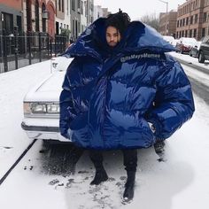 a person in a blue jacket standing next to a car covered in snow on a city street