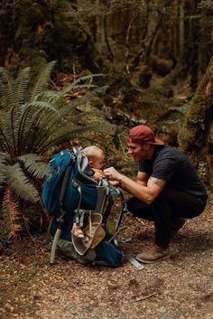 a man kneeling down next to a baby in a backpack on a forest trail with ferns