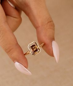 a woman's hand holding a gold ring with a brown diamond on it and white nails